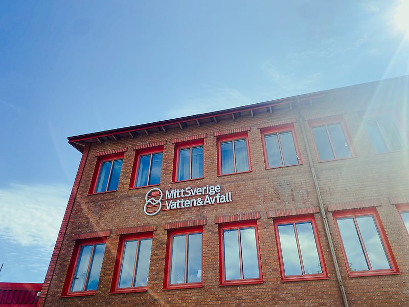 Rött tegelhus mot blå himmel med en skylt med logotypen MittSverige Vatten & Avfall, foto.