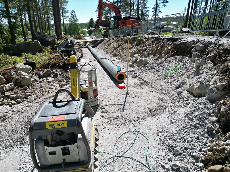 Bild på ledning som grävs ner i marken. I bakgrunden syns en grävmaskin och två yrkesarbetare.
