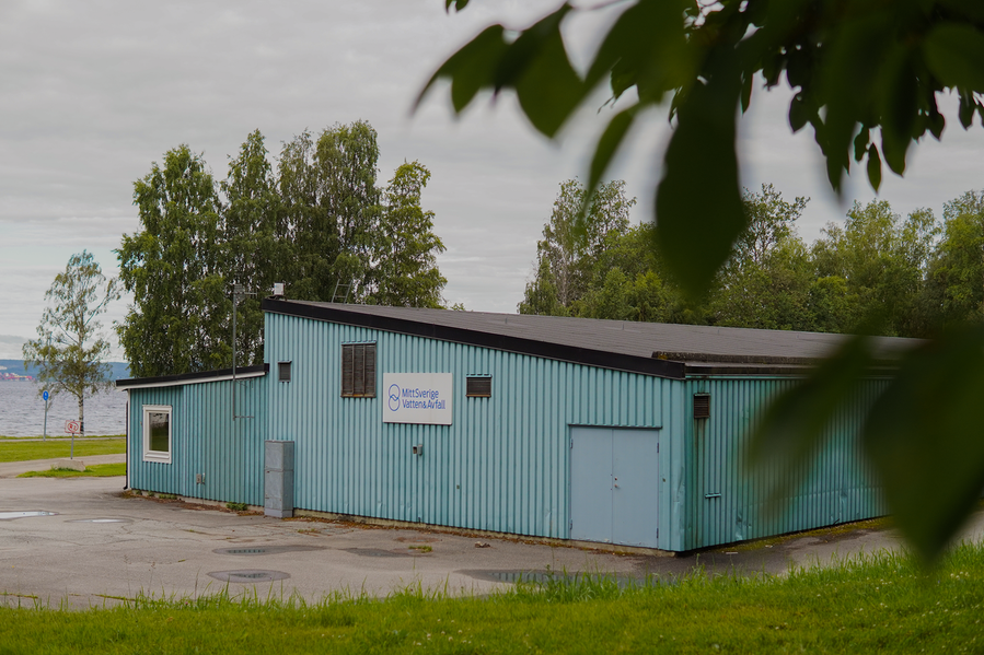 Avloppsreningsverk i Söråker. Husbyggnaden är blå med en MittSverige Vatten och Avfalls skylt på fasaden. Gräset och träden är gröna, himlen är grå och molnig, foto.