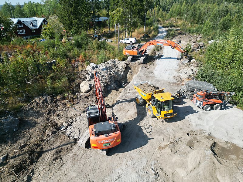 Grävmaskiner som arbetar med att lasta massor på en dumper längs en landsväg, solen skiner och träden är gröna, foto.